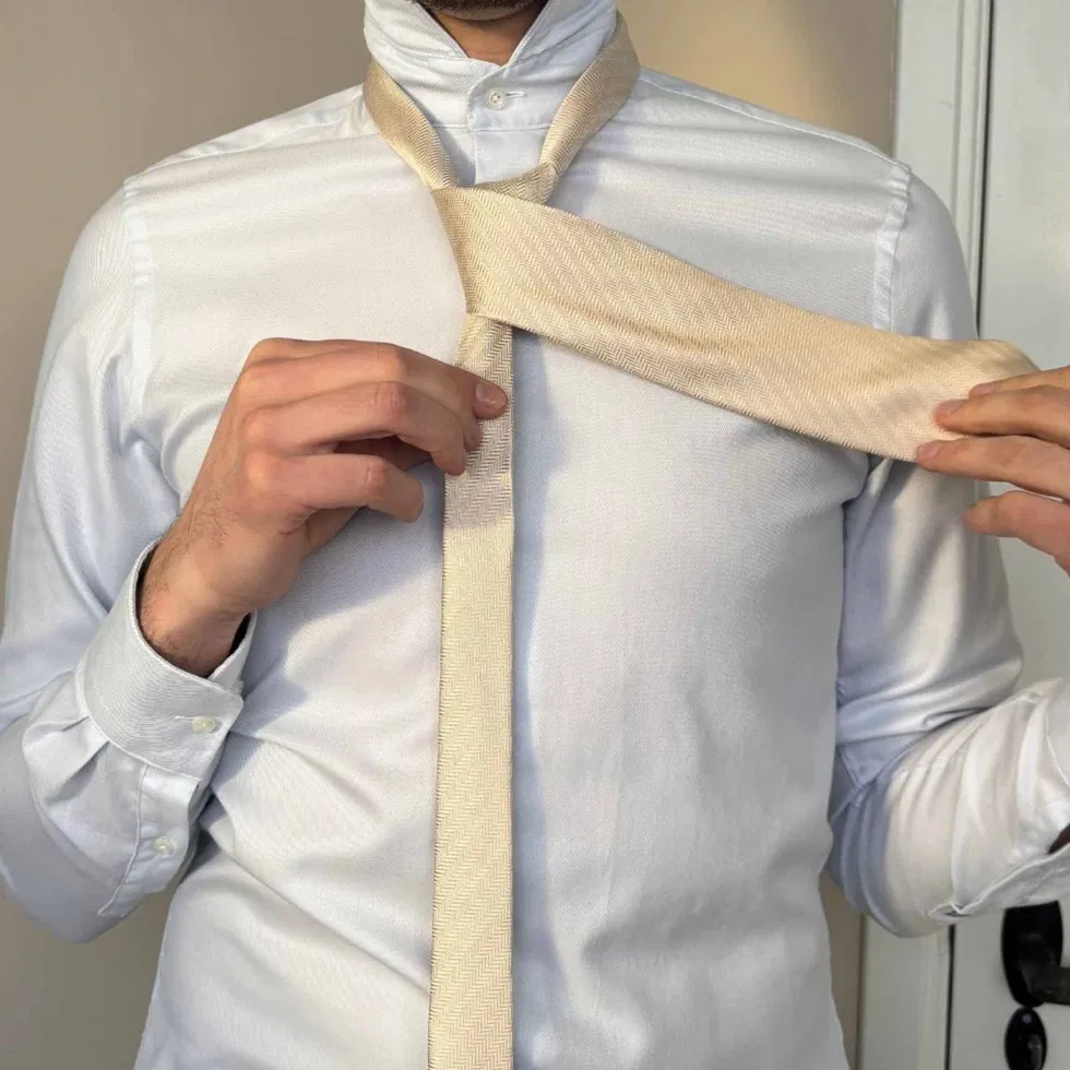 Die breite Seite der Krawatte wird über die schmale Seite nach links gelegt, um den Knoten zu formen.