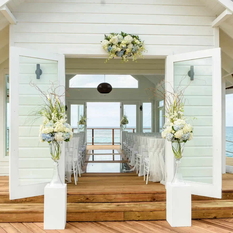 Geöffnete Türen einer weiß gestrichenen Strandkapelle führen zu einem mit Blumen geschmückten Innenraum, dahinter das weite Meer.