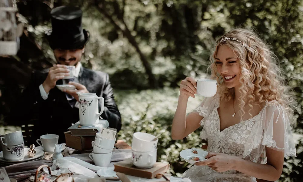 Ein Brautpaar im Alice im Wunderland-Stil sitzt lachend an einem Tisch im Freien und genießt eine Tasse Tee. Die Braut trägt ein Spitzenkleid und der Bräutigam einen Zylinder.