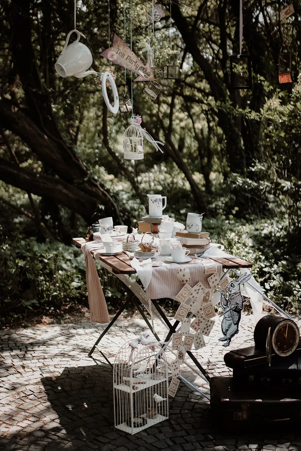 Ein reich dekorierter Tisch im Wald, auf dem viele Tassen, Teller und Bücher verteilt sind. Über dem Tisch hängen Teekannen und Vogelkäfige in der Luft.