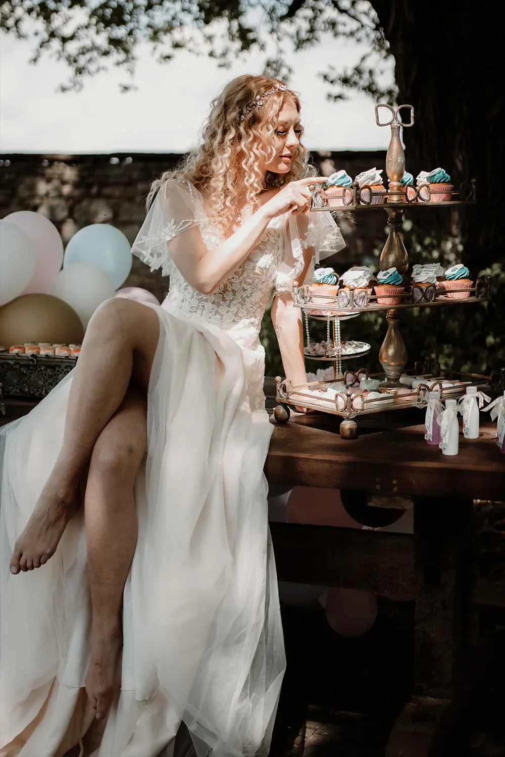 Eine Braut in einem Spitzenkleid sitzt neben einem dreistufigen Cupcake-Ständer und wählt einen kunstvoll dekorierten Cupcake aus.