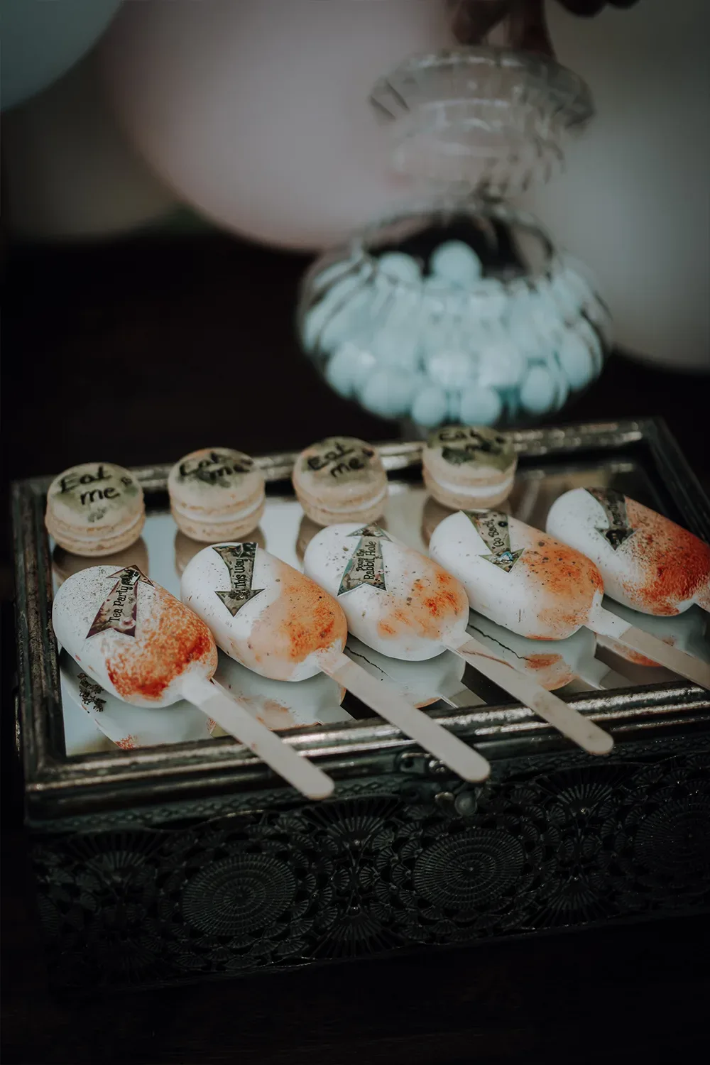 Mini-Desserts und Macarons mit der Aufschrift "Eat Me" auf einem silbernen Tablett, passend zum "Alice im Wunderland"-Thema.