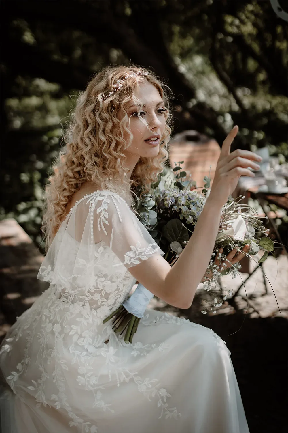 Eine Braut mit lockigem, blonden Haar und einem eleganten Blumenkranz in ihrem Haar zeigt auf etwas und trägt einen üppigen Brautstrauß.