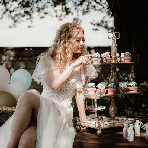 Die Braut, gekleidet in einem romantischen weißen Spitzenkleid, sitzt neben einem Etagere mit dekorierten Cupcakes und betrachtet liebevoll die süßen Leckereien. Im Hintergrund sind bunte Luftballons zu sehen.