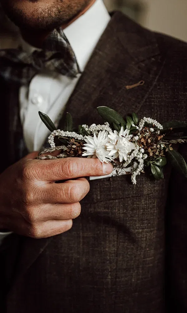 Ein Mann im stilvollen Anzug richtet seinen floralen Anstecker, der aus weißen Blüten und zarten grünen Akzenten besteht, an seinem Revers.