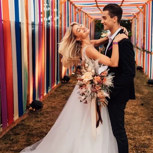 Ein Brautpaar tanzt in einem farbenfrohen Gang, der mit bunten Bändern dekoriert ist. Die Braut trägt ein elegantes weißes Kleid und hält einen Blumenstrauß, während der Bräutigam in einem schwarzen Anzug sie zärtlich umarmt.