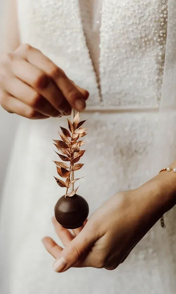Die Braut hält einen eleganten Cake-Pop mit einer goldenen, filigranen Dekoration als Teil des Hochzeitsdesserts.