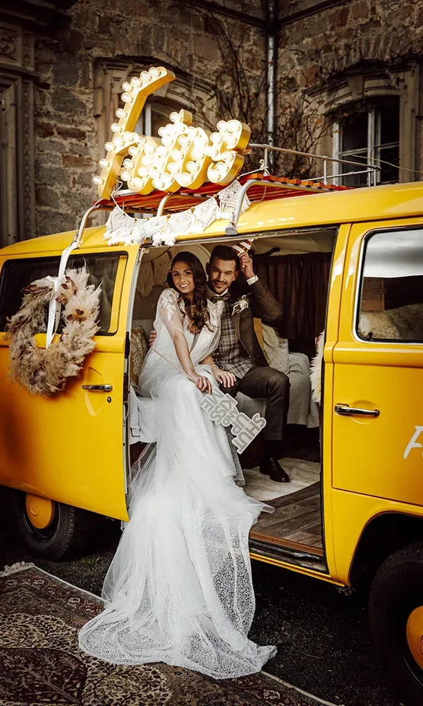 Das Brautpaar sitzt glücklich in einem gelben VW-Bus, der mit einem „Fotos“-Schild dekoriert ist. Die Braut trägt ein weißes Kleid mit Schleier.