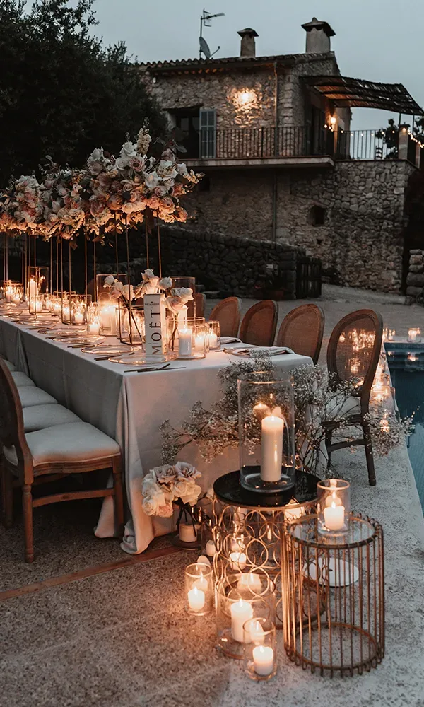 Eine elegante Abend-Tischdekoration unter freiem Himmel, mit vielen brennenden Kerzen und Blumenarrangements vor einem steinernen Gebäude.