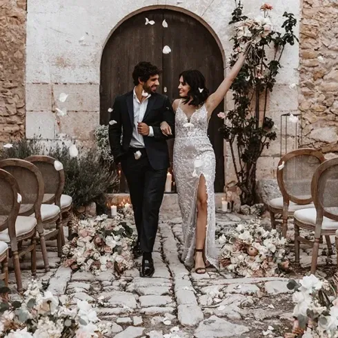 Ein Brautpaar läuft fröhlich durch einen mit Rosenblättern bestreuten Gang nach der Trauung, die Braut trägt ein elegantes Kleid mit Schlitz.