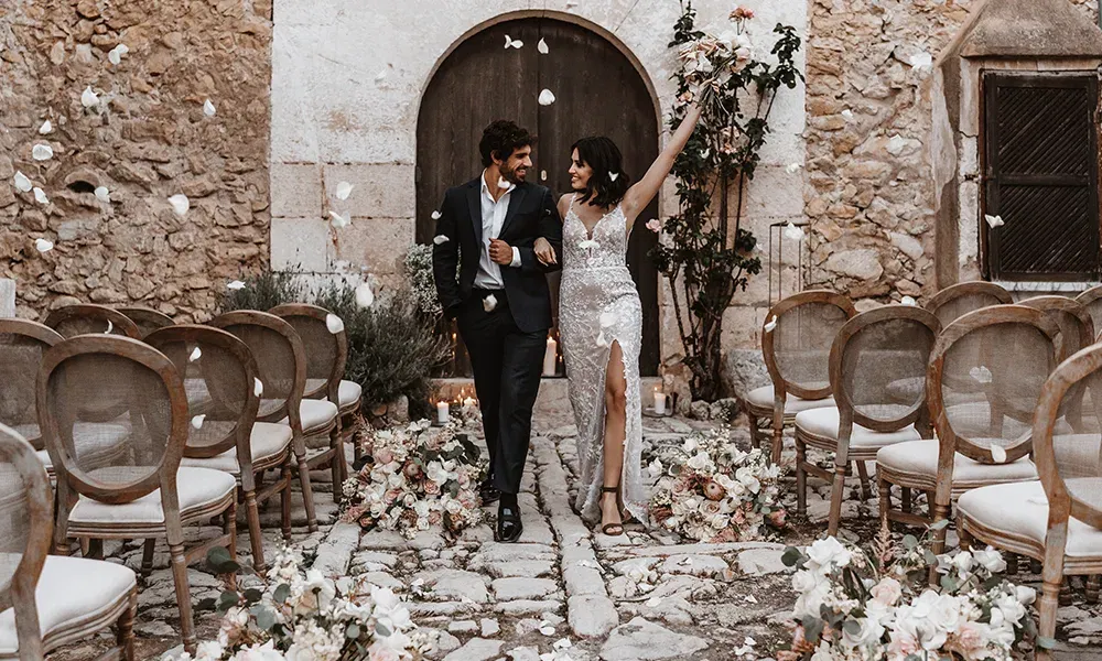 Ein Brautpaar läuft fröhlich durch einen mit Rosenblättern bestreuten Gang nach der Trauung, die Braut trägt ein elegantes Kleid mit Schlitz.