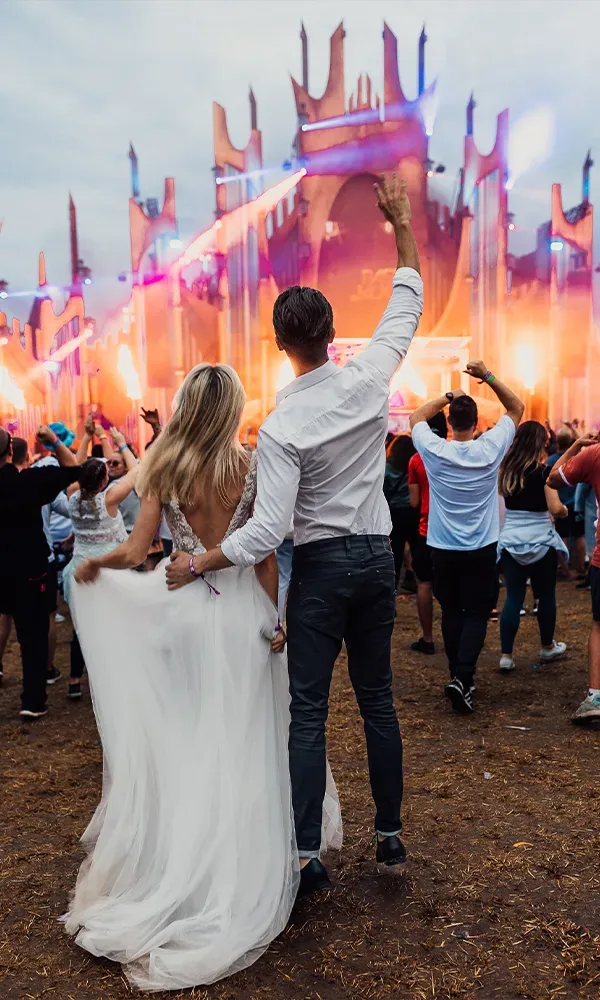 Ein Hochzeitspaar von hinten, das auf einem Festival vor einer großen, beleuchteten Bühne steht. Die Braut trägt ein langes, weißes Kleid, während der Bräutigam einen weißen Hemd und dunkle Hose anhat.