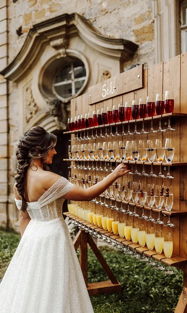 Eine Braut in einem schulterfreien Kleid greift ein Glas Sekt von einer rustikalen Holz-Sektwand.