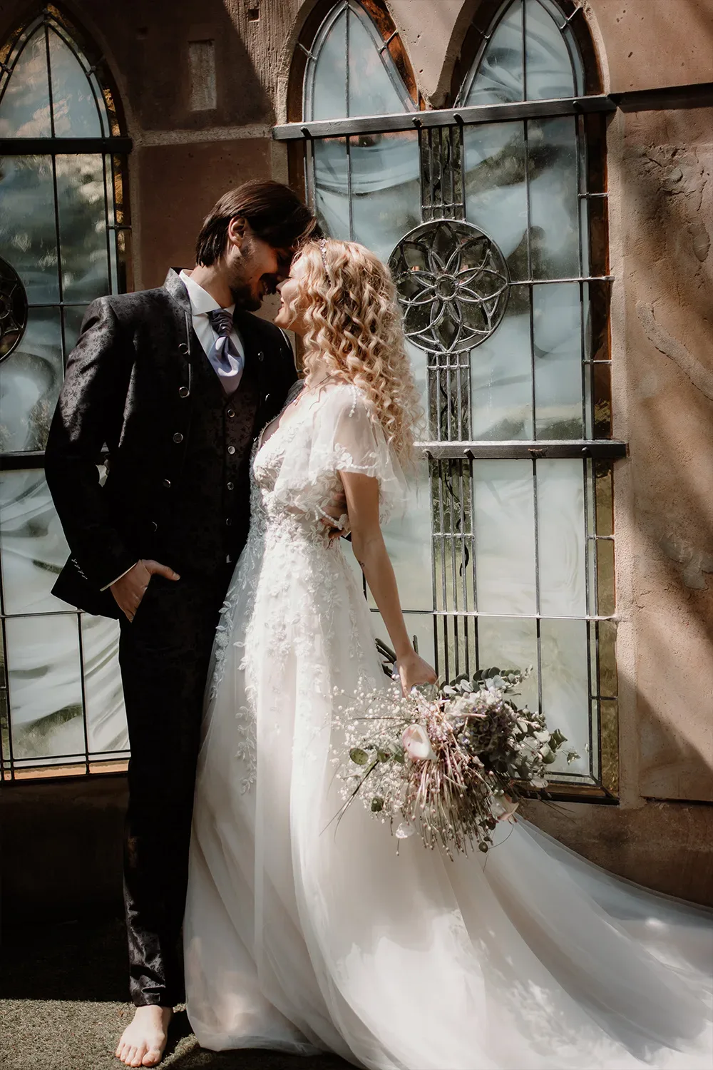Ein romantisches Bild eines Hochzeitspaares. Die Braut in einem weißen, Spitzenkleid lehnt sich liebevoll an ihren Bräutigam, der in einem eleganten schwarzen Anzug steht. Im Hintergrund sind bunte Fenster zu sehen, die das Bild mit weichem Licht durchfluten.