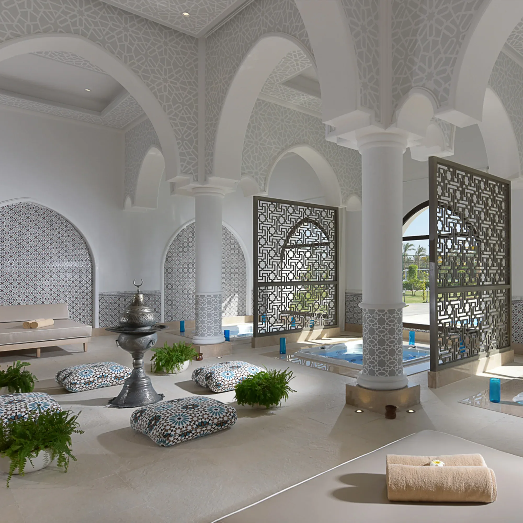 Ein luxuriöses Spa mit arabisch inspiriertem Design, das durch filigrane Gitter, Säulen und Mosaikmuster beeindruckt. Im Raum befinden sich niedrige, gemusterte Sitzkissen, Grünpflanzen und ein dekoratives Wasserbecken.