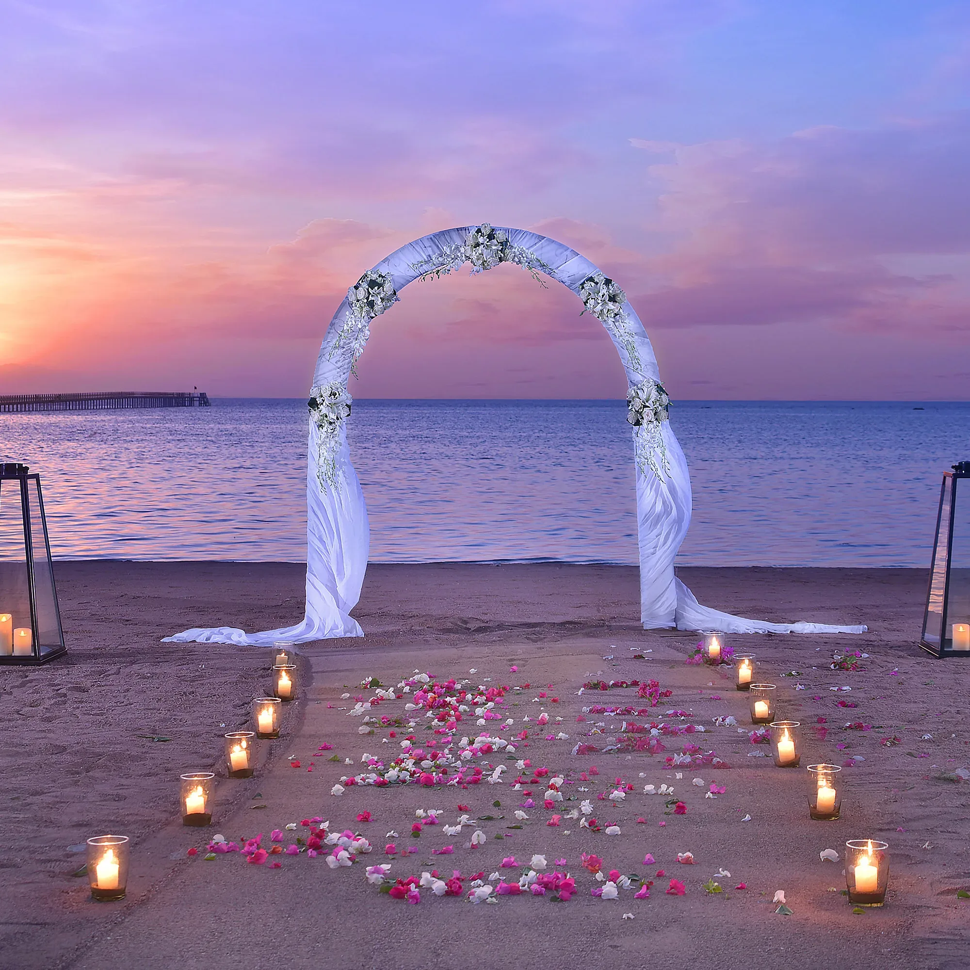 Ein romantisches Strand-Setup bei Sonnenuntergang, mit einem mit Blumen verzierten Hochzeitsbogen. Der Gang ist mit Blütenblättern und Kerzen gesäumt, während die ruhige See im Hintergrund glitzert.