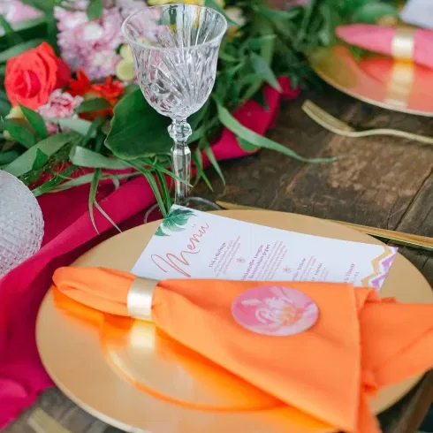 Ein festlich gedeckter Tisch mit einer orangefarbenen Serviette, die auf einem goldfarbenen Teller liegt, umgeben von einem eleganten Kristallglas und einem farbenfrohen Menü, das die Tischdekoration ergänzt.
