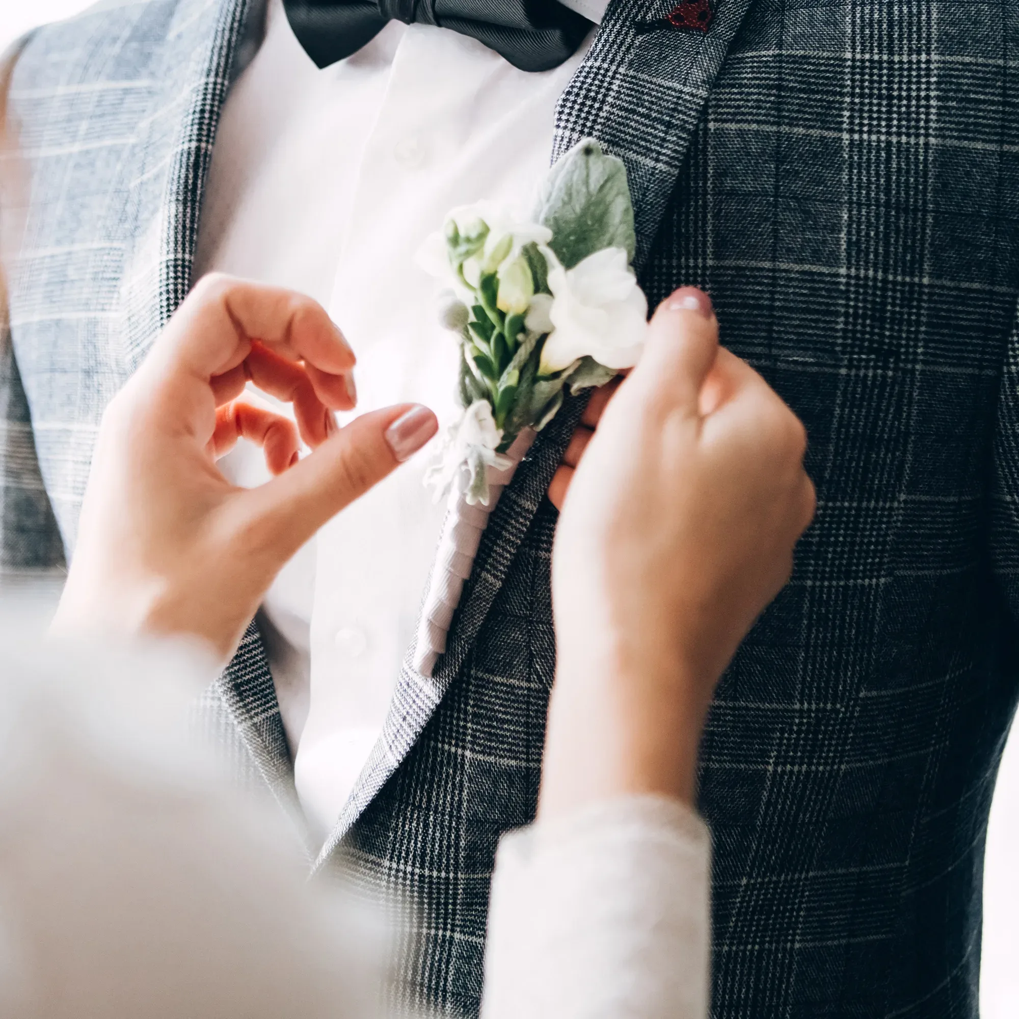 Eine Nahaufnahme eines Bräutigams in einem grauen Karoanzug. Jemand befestigt eine weiße Blumen-Boutonnière an seinem Revers, während er eine schwarze Fliege trägt.