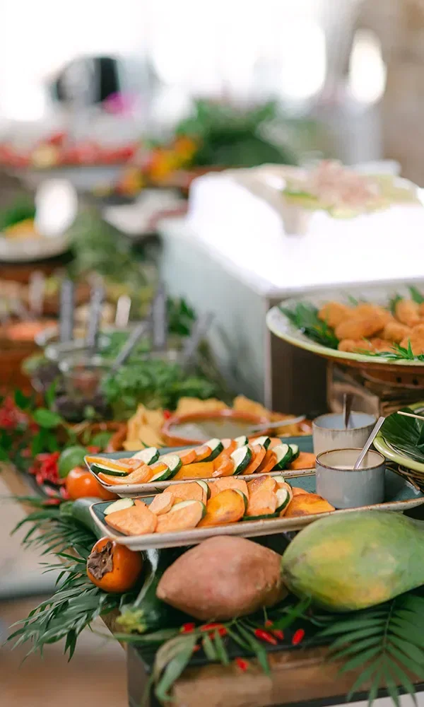 Ein üppiges Hochzeitsbuffet mit einer Vielfalt an bunten Gerichten. Auf dem vorderen Tablett befinden sich gebackene Zucchinischeiben und Süßkartoffeln, umgeben von tropischen Früchten und Gemüse.