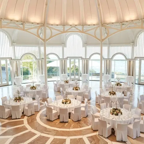 Ein eleganter, heller Bankettsaal mit hoher Decke und großen Fenstern, in dem mehrere runde Tische mit weißen Stühlen für eine Hochzeit eingedeckt sind. Die Tische sind mit Blumenarrangements dekoriert und bieten einen Blick auf die umliegende Landschaft.
