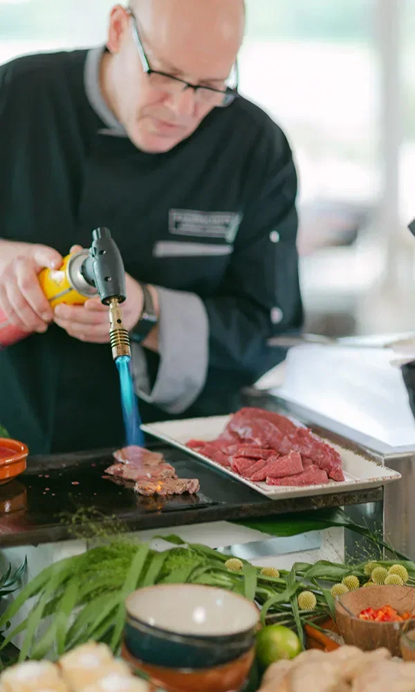 Ein Koch in schwarzer Kochuniform flambiert ein Stück Fleisch auf einer heißen Platte mit einem Flambierbrenner, umgeben von frischen Kräutern und Zutaten.