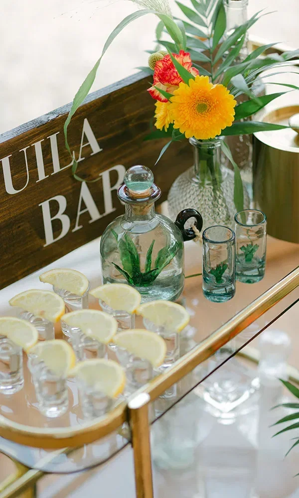 Eine stilvolle Tequila-Bar mit einer Flasche Tequila, Shotgläsern und Zitronenscheiben auf einem goldenen Tablett. Bunte Blumen und dekorative Vasen ergänzen das tropische Ambiente.