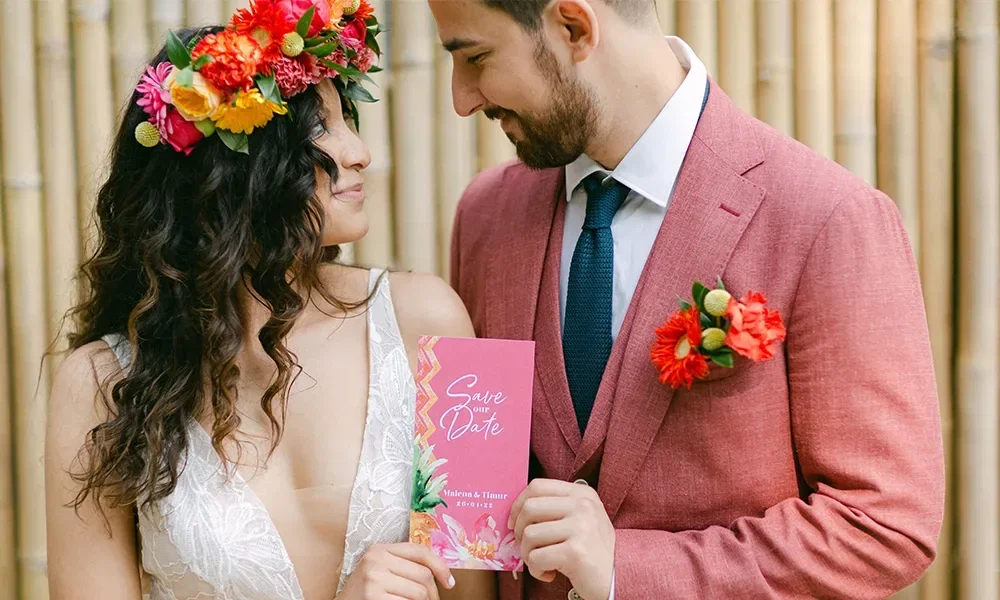 Ein glückliches Paar, sie trägt einen Blumenkranz im Haar und er einen eleganten Anzug in Rosa. Gemeinsam halten sie eine rosafarbene "Save the Date" Karte vor sich und lächeln sich liebevoll an.