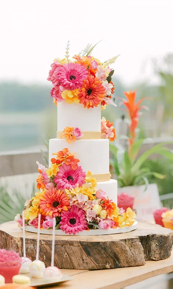 Eine mehrstöckige Hochzeitstorte, verziert mit einer Fülle von bunten Blumen in Rosa, Gelb und Orange. Die Torte steht auf einem rustikalen Holztisch und strahlt sommerliche Frische aus.