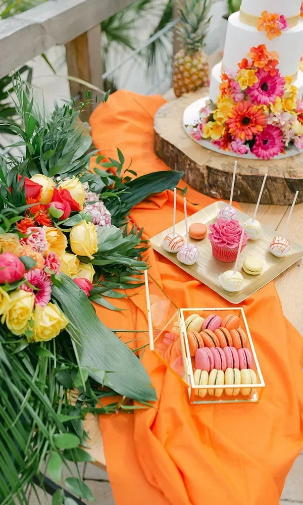 Ein dekorativer Sweet Table mit bunten Macarons, Cupcakes und einer tropisch dekorierten Hochzeitstorte. Um den Tisch herum liegen bunte Blumen und grüne Blätter.