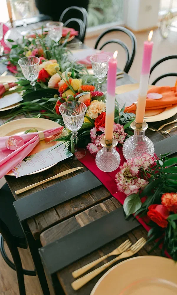 Eine festliche Tischdekoration mit bunten Blumen, eleganten Kristallgläsern und brennenden rosa Kerzen, die einen lebendigen Kontrast zu den goldenen Tellern und Servietten bieten.
