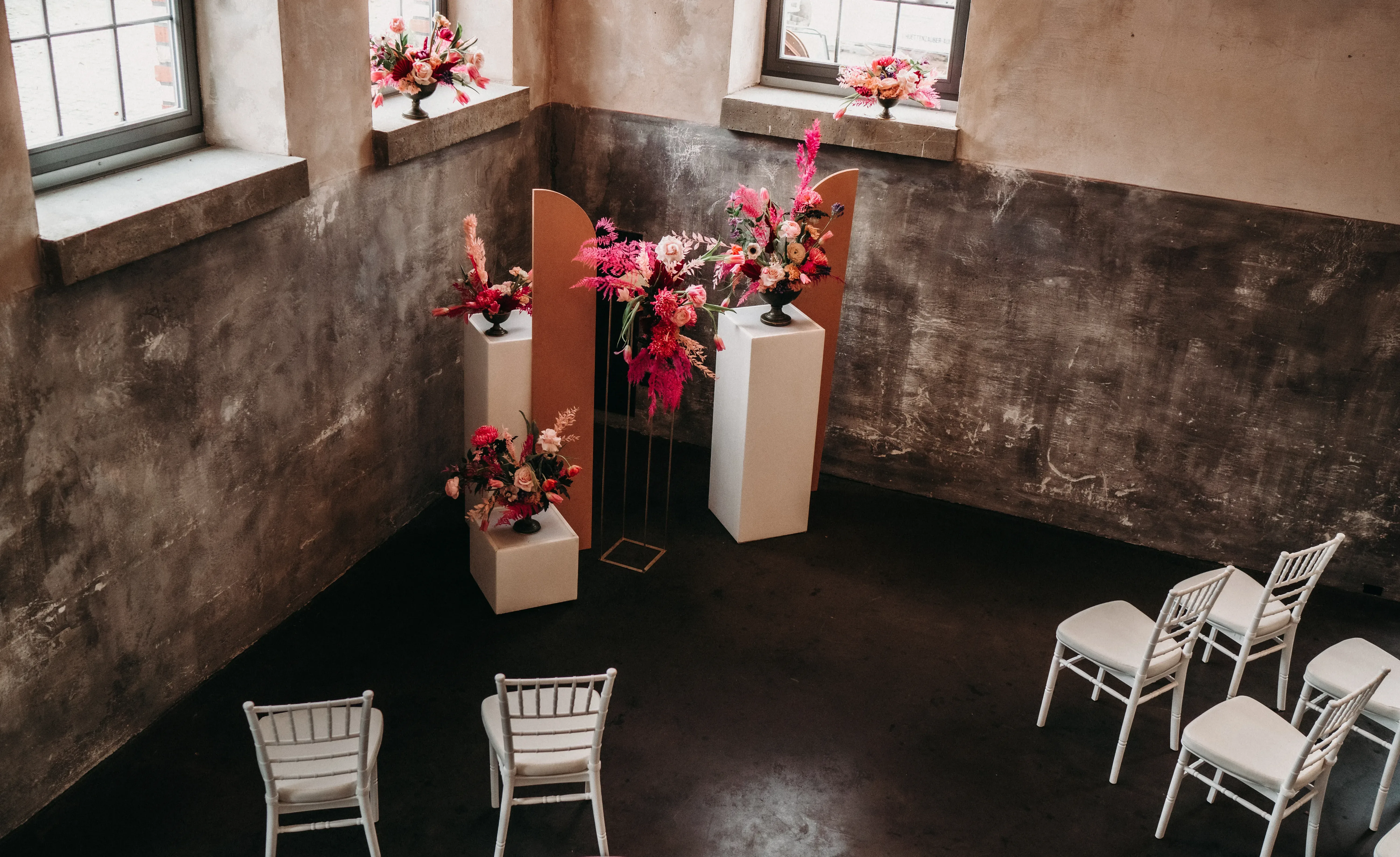 Eine rustikale Eventlocation mit alten Steinwänden und großen Fenstern. Weiße Stühle stehen im Halbkreis um einen zentralen Bereich mit floralen Dekorationen in Rosa- und Rottönen, die auf hohen Sockeln platziert sind. Der minimalistisch gestaltete Raum wird von natürlichem Licht durch die Fenster durchflutet.