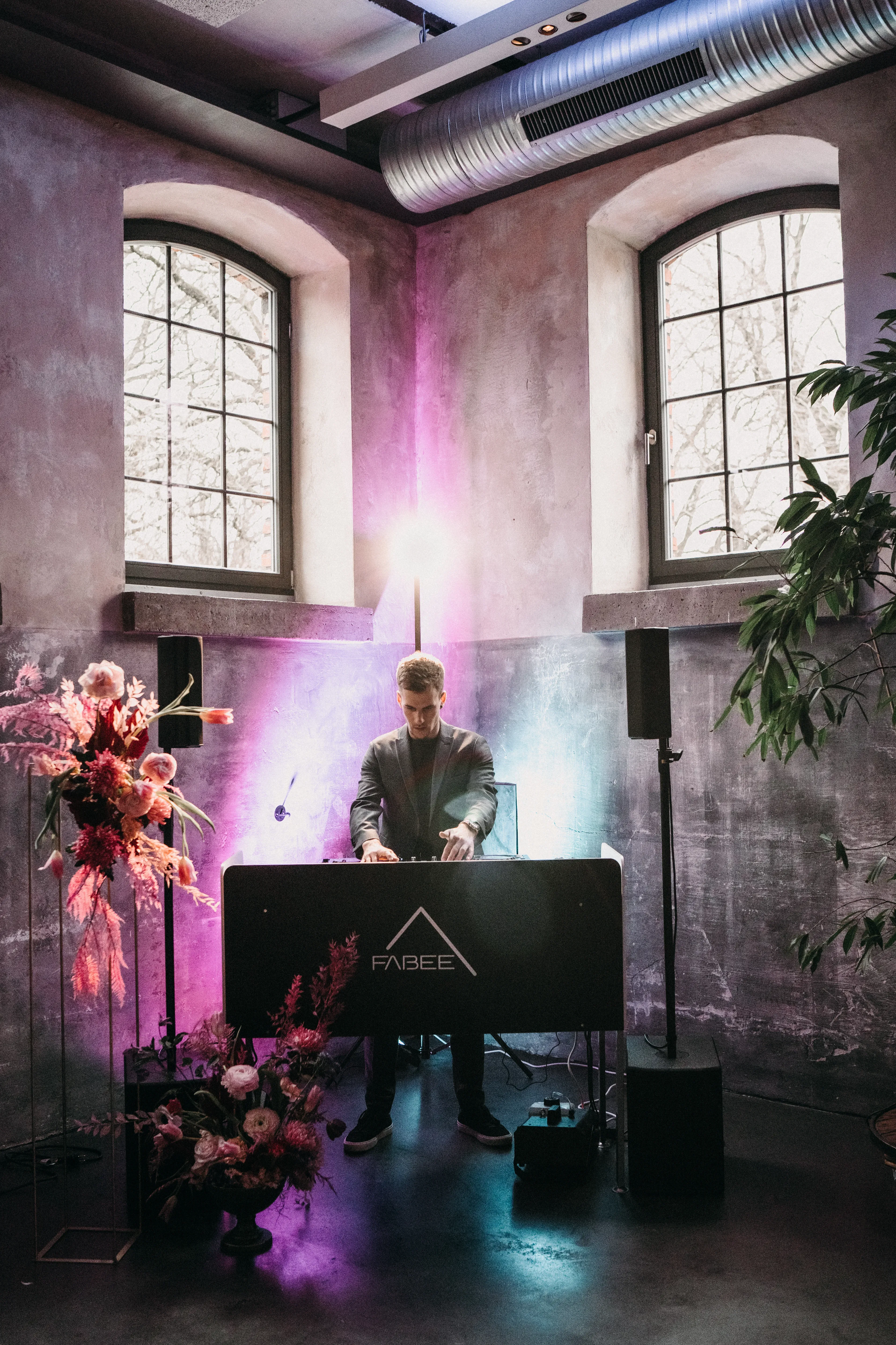 Ein DJ performt in einem stilvollen Raum mit hohen Fenstern und sichtbar freiliegenden Lüftungsrohren an der Decke. Bunte Scheinwerfer in Lila und Blau leuchten den Raum aus, während auf der linken Seite Blumenarrangements mit kräftigen Rosa- und Rottönen den Raum schmücken.