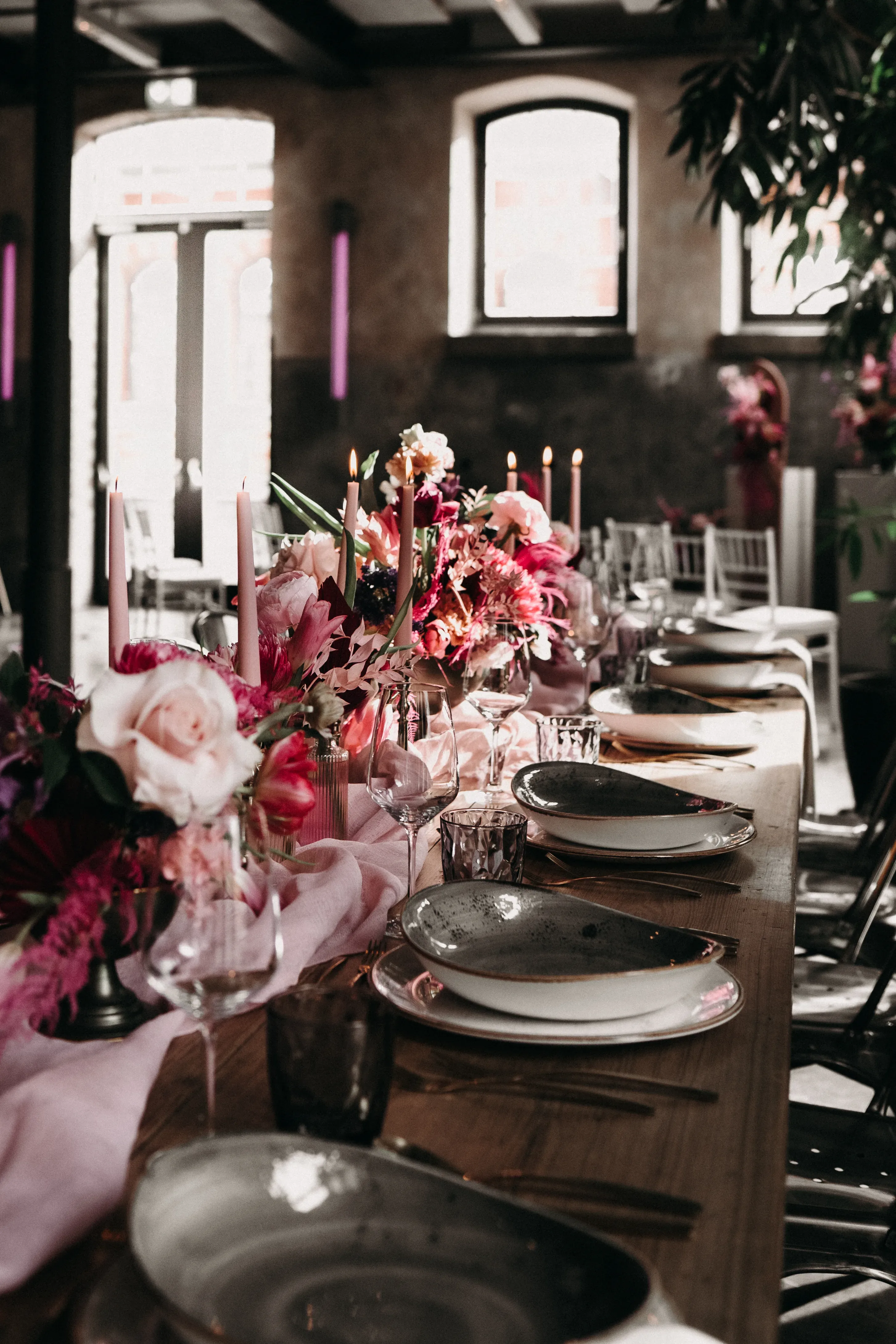 Eine festlich gedeckte Hochzeitstafel mit elegant arrangierten Tellern und Gläsern, dekoriert mit üppigen Blumenarrangements in Rosa- und Pinktönen sowie hohen, brennenden Kerzen. Der Tisch ist mit einer sanften Stoffbahn dekoriert, die sich durch das gesamte Gedeck zieht, während Sonnenlicht durch die Fenster fällt und den Raum erhellt.