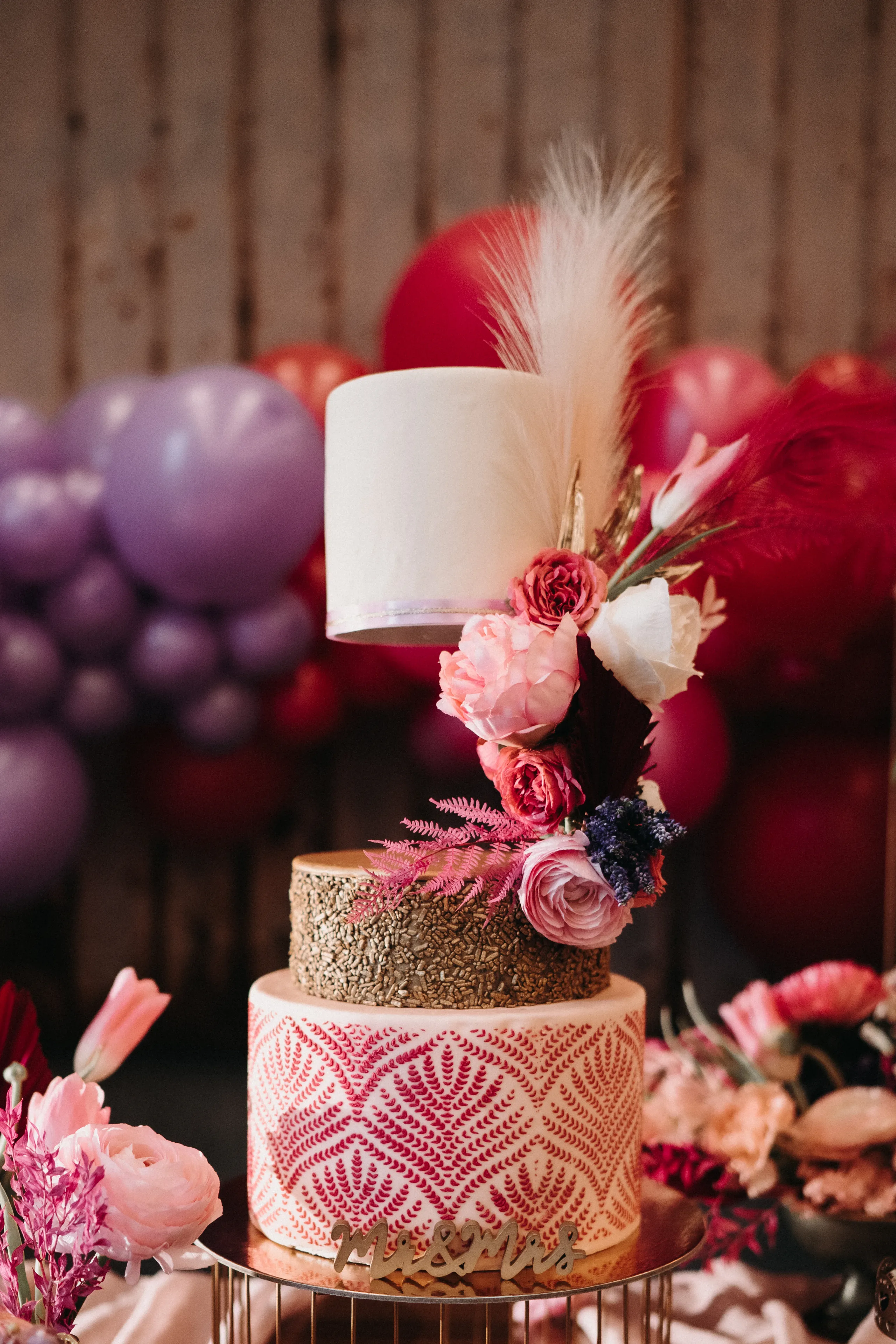 Eine dreistufige Hochzeitstorte, kunstvoll dekoriert mit floralen Elementen und pinken Blüten, steht im Vordergrund. Die oberste Schicht der Torte ist schlicht weiß, während die mittlere Schicht mit goldenen Texturen verziert ist und die unterste Schicht ein aufwendiges pink-weißes Muster zeigt. Im Hintergrund sind lilafarbene und rote Luftballons dekoriert.