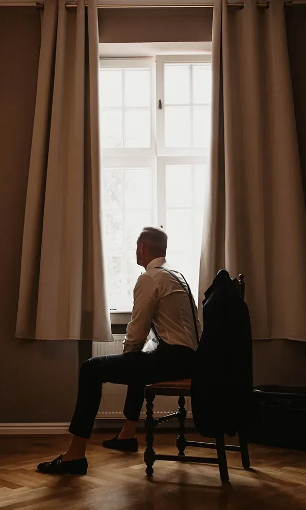 Ein Bräutigam sitzt auf einem Stuhl vor einem großen, lichtdurchfluteten Fenster. Er trägt Hemd und Hosenträger und blickt nachdenklich nach draußen. Sein Jackett hängt über dem Stuhl, während der Raum in warmem Licht getaucht ist.