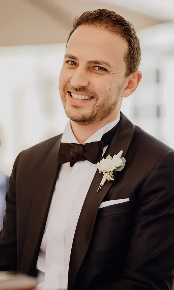 Ein lächelnder Bräutigam in einem eleganten schwarzen Anzug mit Fliege und einer weißen Blume im Knopfloch. Er wirkt entspannt und glücklich.