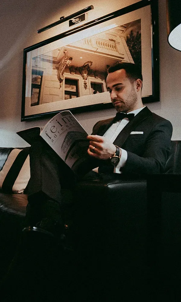 Ein Bräutigam sitzt in einem eleganten schwarzen Anzug auf einem Ledersofa und liest ein Magazin. Im Hintergrund hängt ein gerahmtes Bild, während die gedämpfte Beleuchtung eine entspannte Atmosphäre schafft.