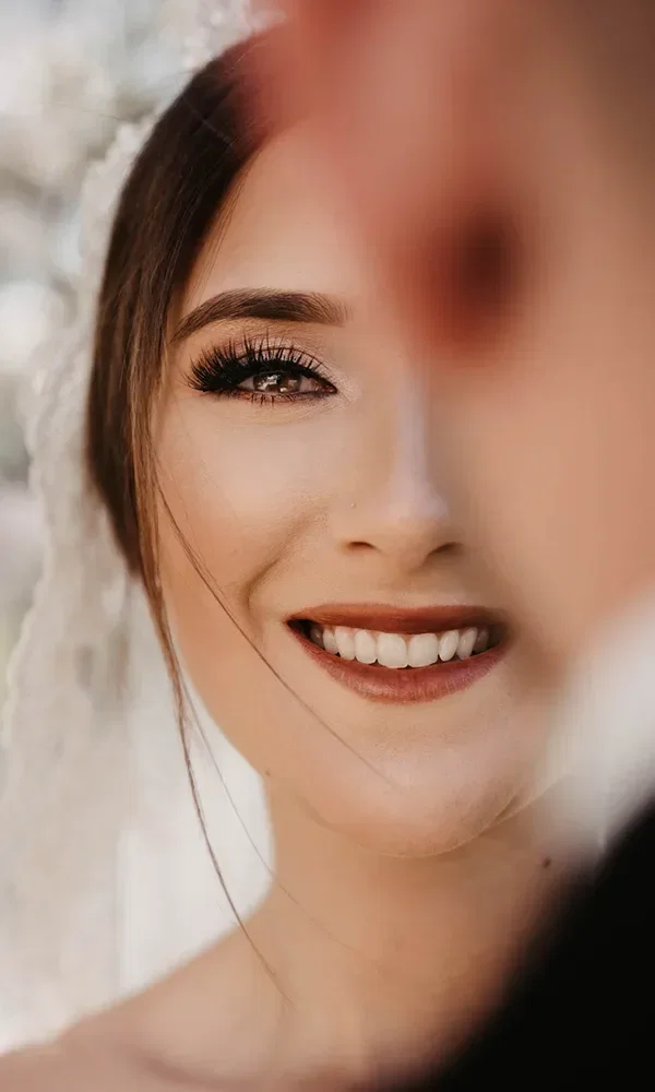 Nahaufnahme des Gesichts einer Braut, die ein breites Lächeln zeigt. Ihr Gesicht wird teils von einem anderen unscharfen Gesicht im Vordergrund verdeckt. Sie trägt ein Brautschleier und hat ein elegantes Make-up mit betonten Wimpern und dezentem Lippenstift.