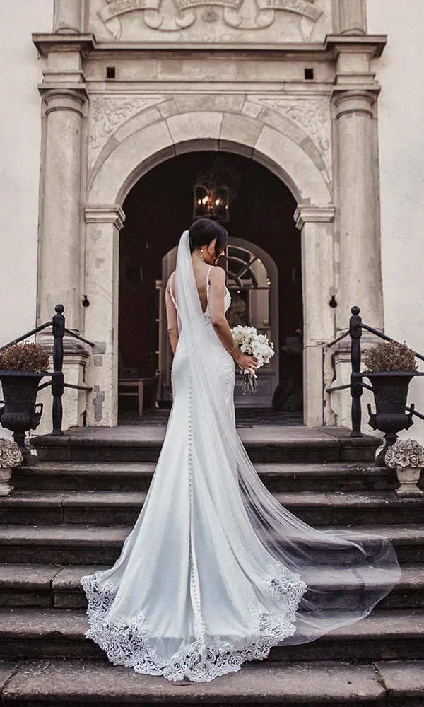 Eine Braut in einem weißen, eleganten Kleid steht mit dem Rücken zur Kamera vor einem großen, verzierten Gebäudeeingang. Ihr langer Schleier fällt über das bodenlange Kleid, und sie hält einen Blumenstrauß in der Hand.