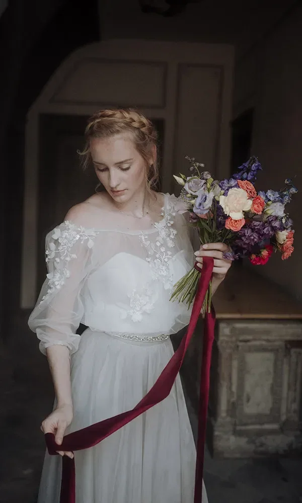 Eine Braut in einem weißen, romantischen Kleid mit floralen Verzierungen hält einen bunten Blumenstrauß und ein rotes Band in der Hand. Sie blickt nach unten und wirkt verträumt.