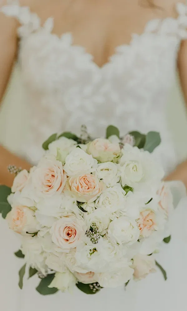 Nahaufnahme eines üppigen Brautstraußes, bestehend aus weißen und zart rosa Rosen, in den Händen einer Braut, die ein weißes Spitzenkleid trägt.