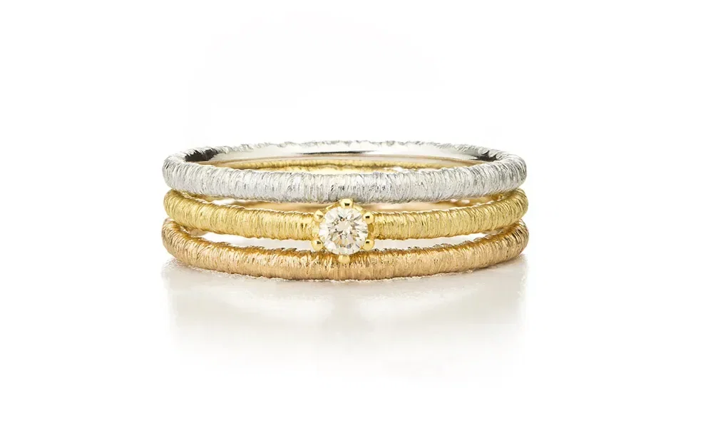 Drei filigrane Stapelringe in verschiedenen Goldtönen. Der mittlere Ring ist mit einem kleinen, funkelnden Diamanten verziert, der die Aufmerksamkeit auf sich zieht. Die Oberfläche der Ringe wirkt strukturiert und handgearbeitet.