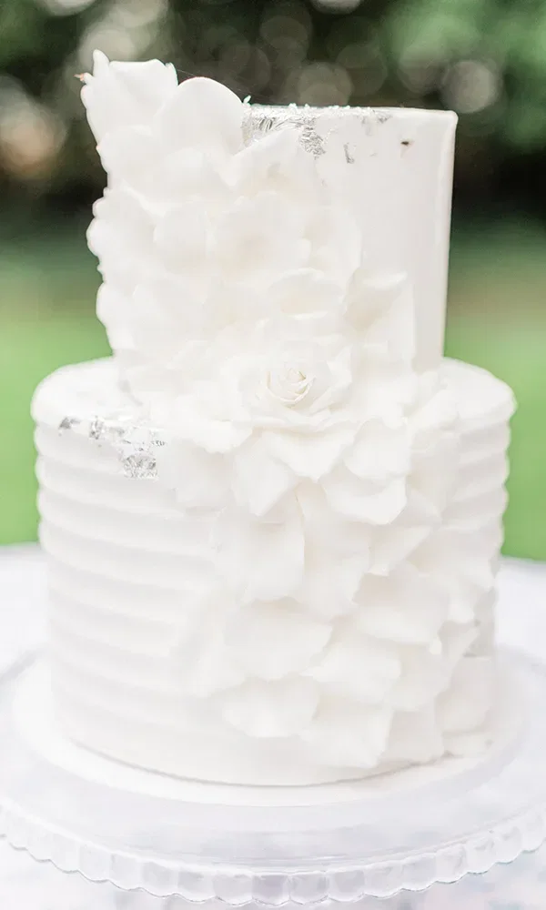 Eine zweistöckige, ganz in Weiß gehaltene Hochzeitstorte mit Blütenverzierungen und dezenten silbernen Akzenten steht auf einem gläsernen Tortenständer im Freien.