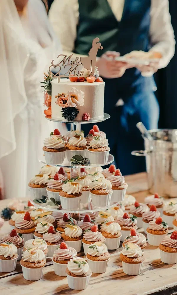 Eine Hochzeitstorte wird von mehreren Cupcakes auf einer mehrstöckigen Etagere umgeben. Die Torte ist mit Beeren und einer "Mr. & Mrs."-Tortenfigur verziert, während das Brautpaar im Hintergrund zu sehen ist.