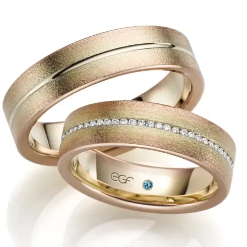 Zwei breite Eheringe aus einer Kombination von Roségold und Gelbgold mit einer matten, strukturierten Oberfläche. Der Ring für die Braut ist mit einer Reihe funkelnder Diamanten verziert, die in die Mitte des Rings eingefasst sind, während der Ring für den Bräutigam eine glatte, polierte Rille aufweist.