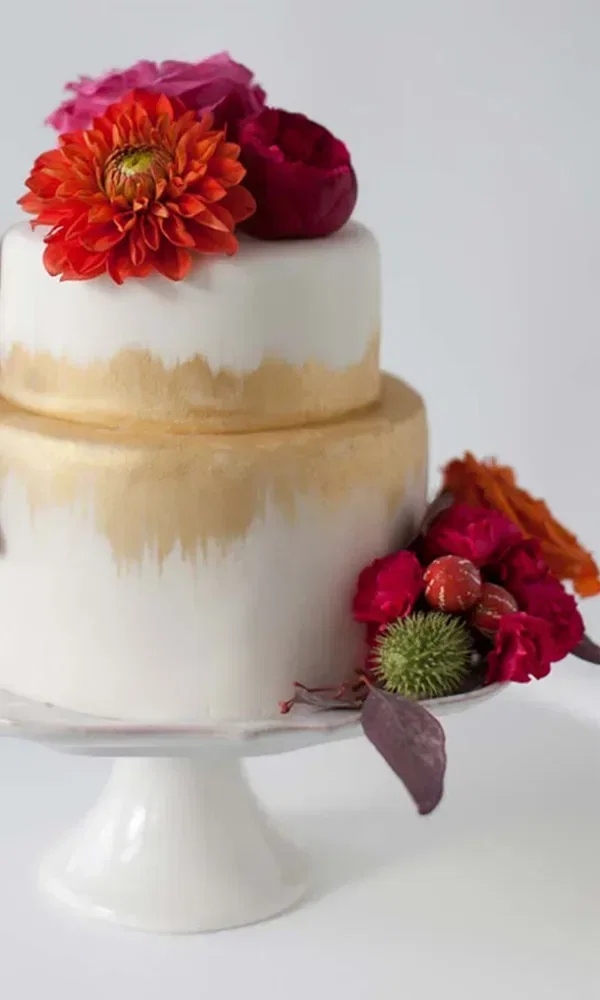 Eine zweistöckige Fondant-Hochzeitstorte mit goldenen Akzenten und bunten, frischen Blumen auf der oberen und unteren Etage steht auf einem weißen Tortenständer.