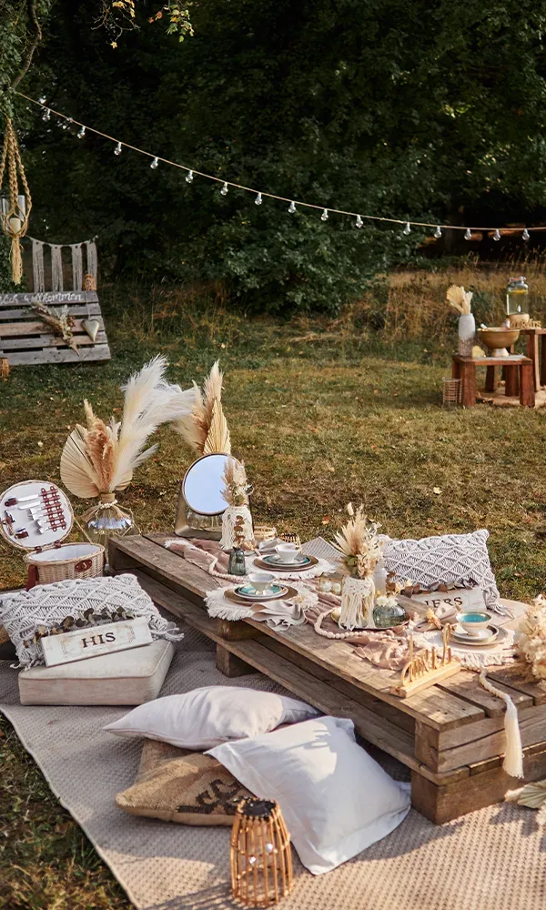 Ein romantisch gedeckter niedriger Holztisch im Freien mit Kissen und Teppichen. Die Tischdekoration umfasst Kerzen, Blumen und Tassen, während eine gemütliche Gartenschaukel und Lichterketten den Hintergrund zieren.