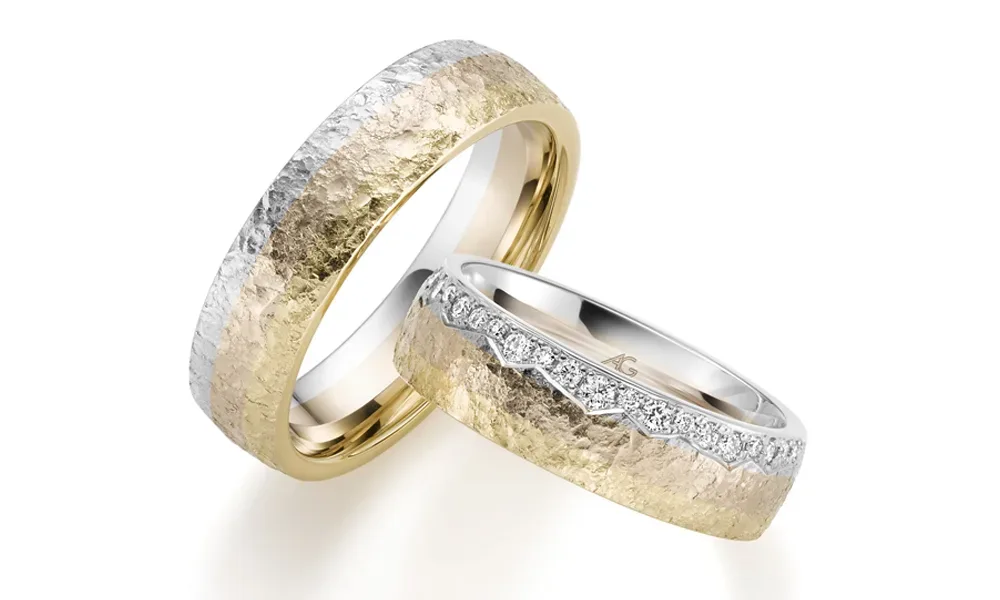 Zwei breite Eheringe aus einer Kombination von Gelb- und Weißgold mit gehämmerter Oberfläche. Der Damenring ist zusätzlich mit einer schimmernden Reihe von Diamanten an der oberen Kante verziert, was ihm eine elegante Note verleiht.