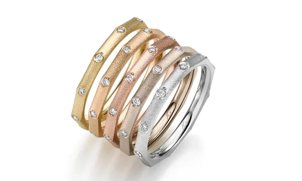 Fünf filigrane Stapelringe in verschiedenen Goldtönen: Gelbgold, Roségold, Weißgold und Platin. Jeder Ring hat eine facettierte Oberfläche und ist gleichmäßig mit funkelnden Diamanten besetzt, die den minimalistischen Stil der Ringe unterstreichen.