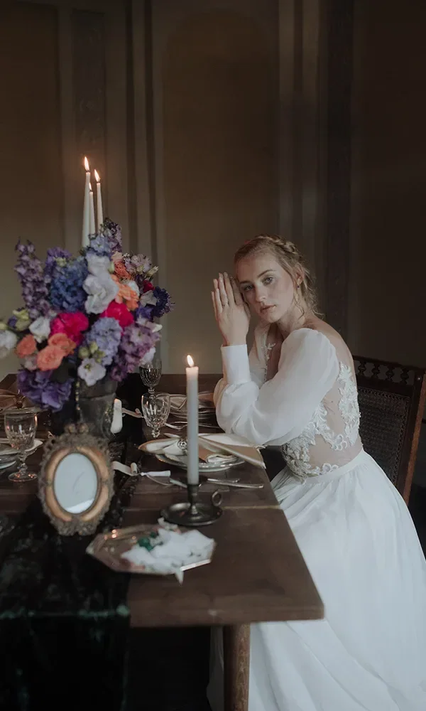Eine Braut in einem weißen Kleid sitzt nachdenklich an einer festlich dekorierten Tafel, auf der brennende Kerzen und ein großer Blumenstrauß mit farbigen Blüten stehen. Sie stützt ihren Kopf auf ihre Hand.